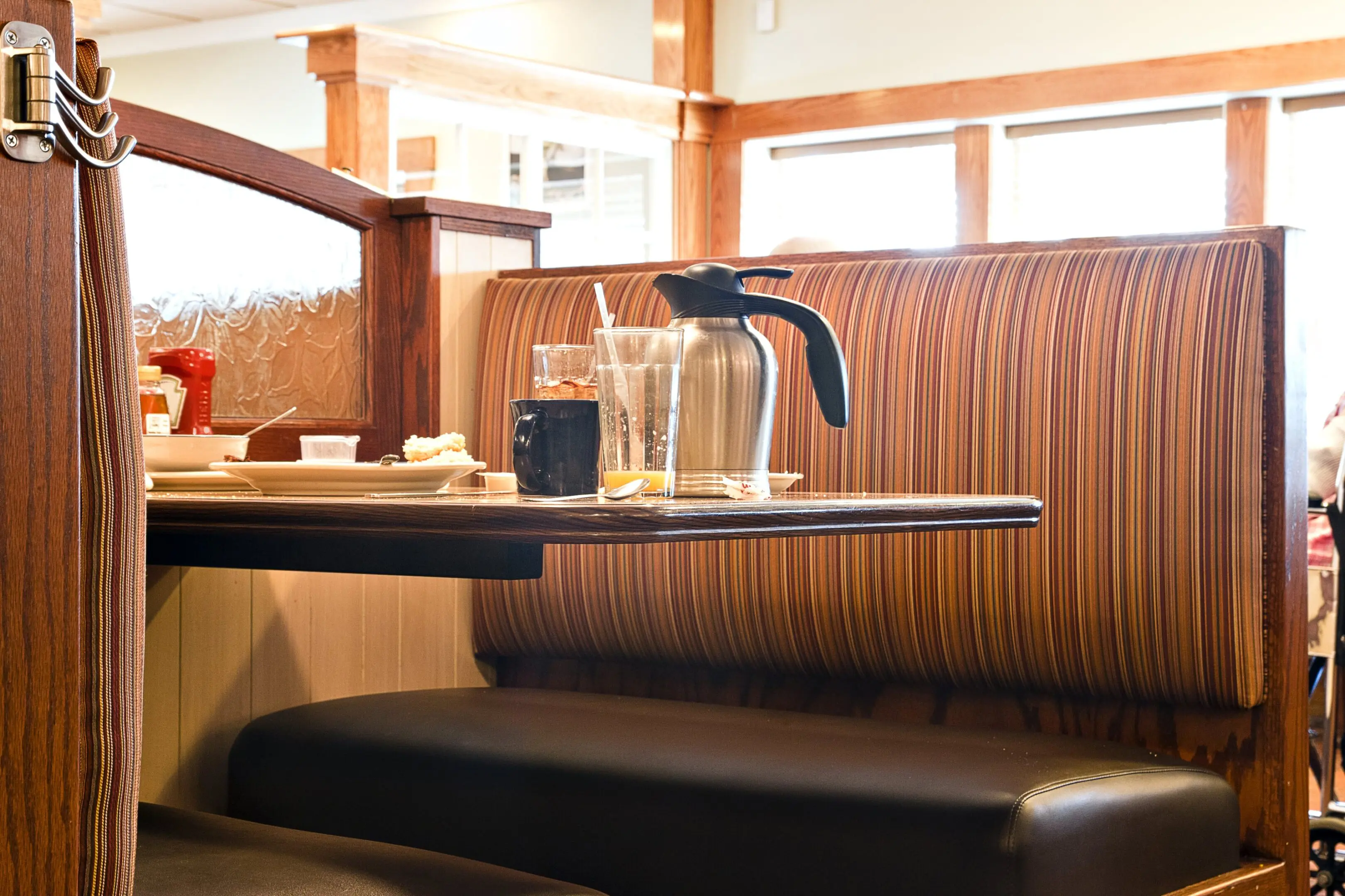 GettyImages-1329257762-min Empty restaurant booth with striped upholstery, dark table, coffee pot, mug, beverage glass, and dining items. Wood paneling and windows in the background.