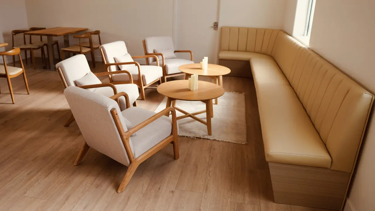 A room with a light beige corner sofa, four wooden armchairs, two round wooden tables, and a row of wooden desks, all on a wooden floor.