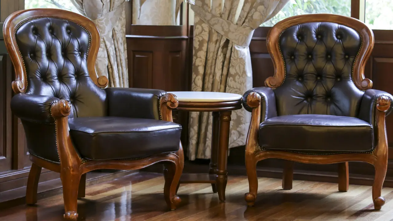 Two black leather armchairs with wooden frames next to a small round table, positioned on a wooden floor in front of curtained windows.
