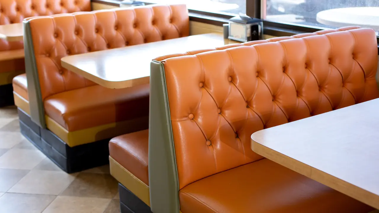 Empty diner booths with orange tufted seats and wooden tables are arranged near large windows, letting in natural light.