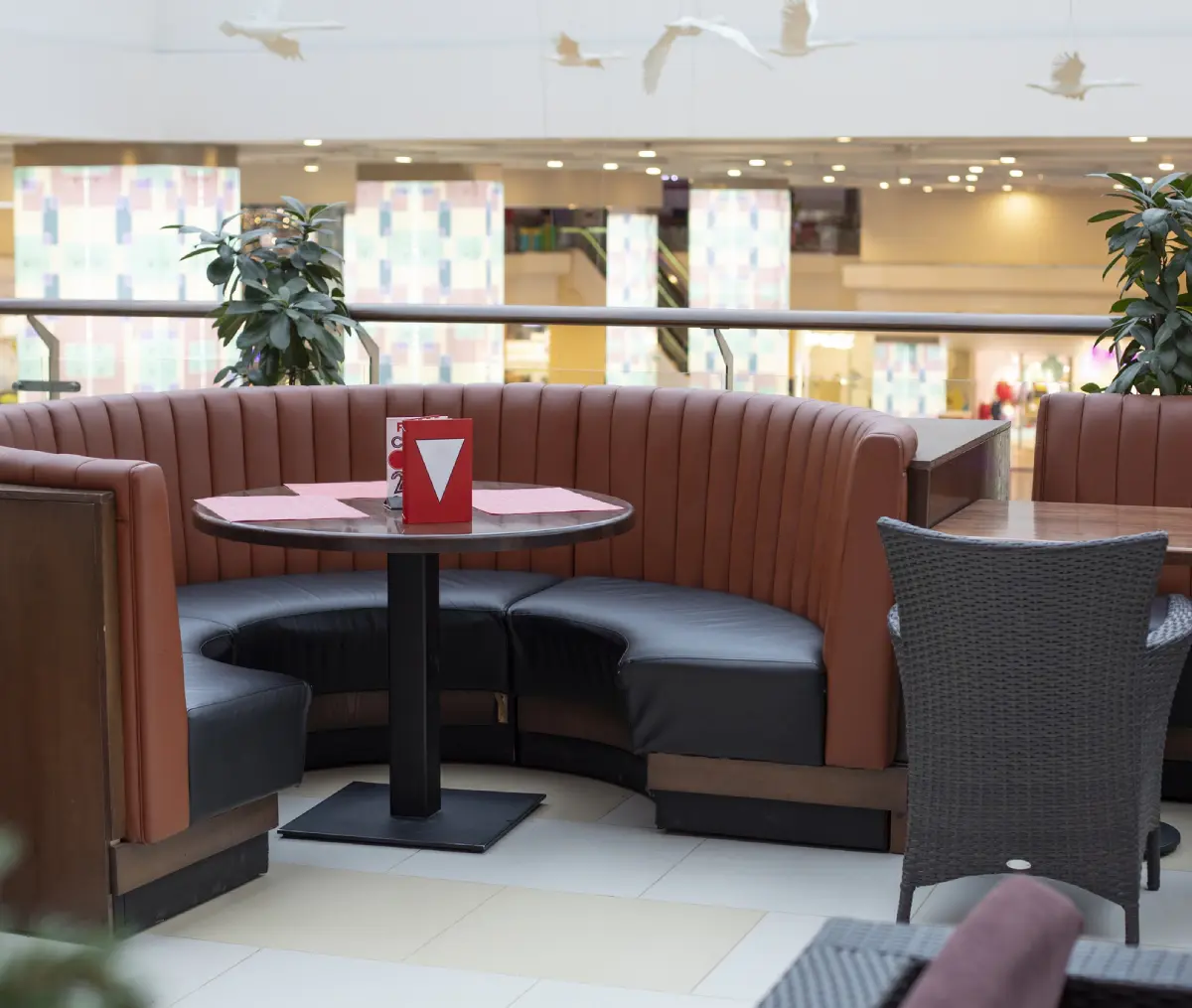 Semi-circular booth with brown leather seating and a round wooden table in a mall. Potted plants and empty chairs are nearby.