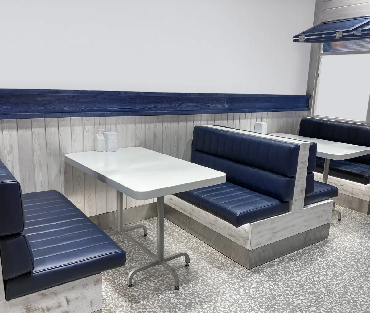 Empty diner booths with blue and white seating and tables, set against a wood-paneled wall, on a speckled floor.
