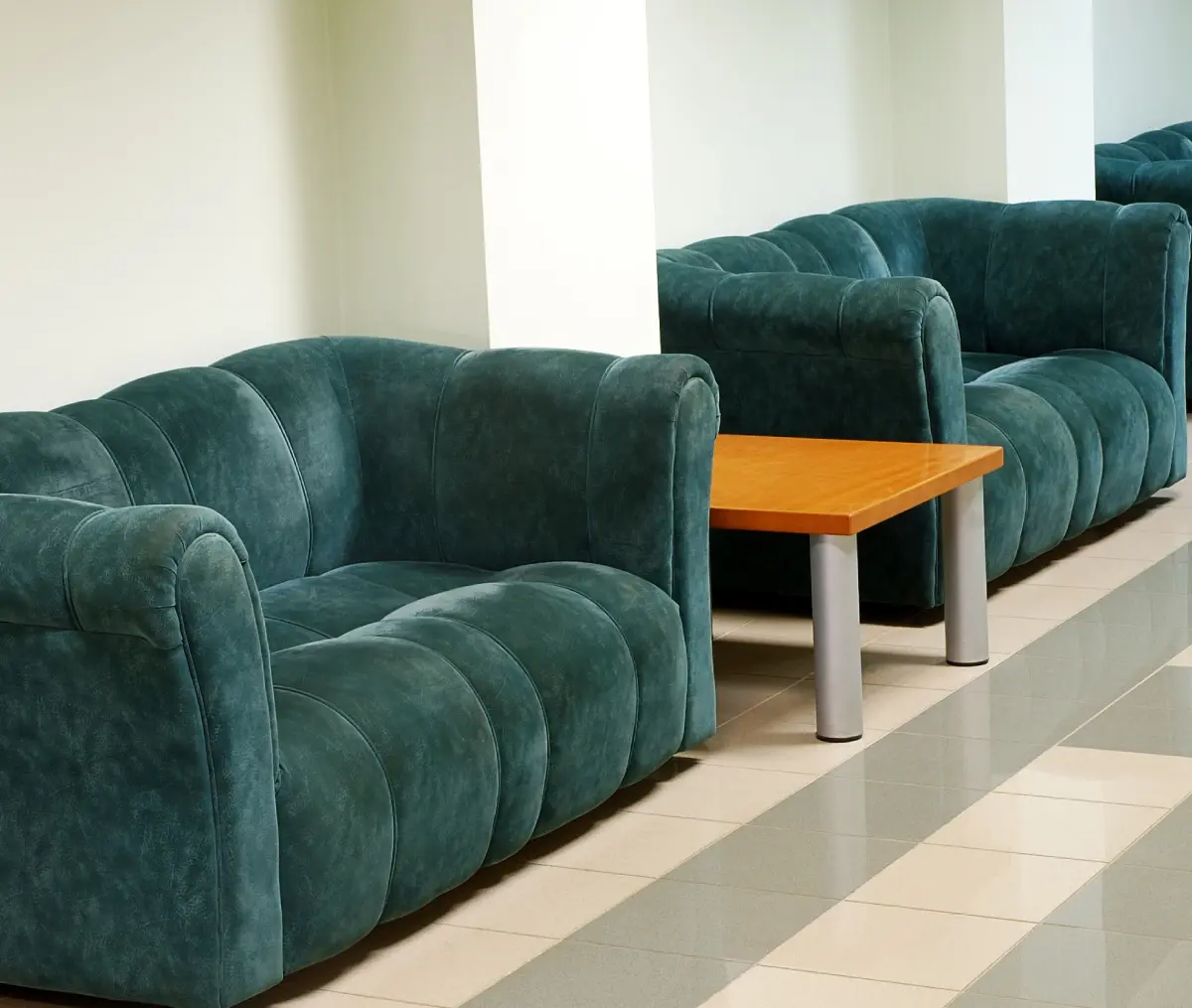 A row of green sofas with a wooden coffee table in between, placed along a tiled hallway with cream-colored walls.