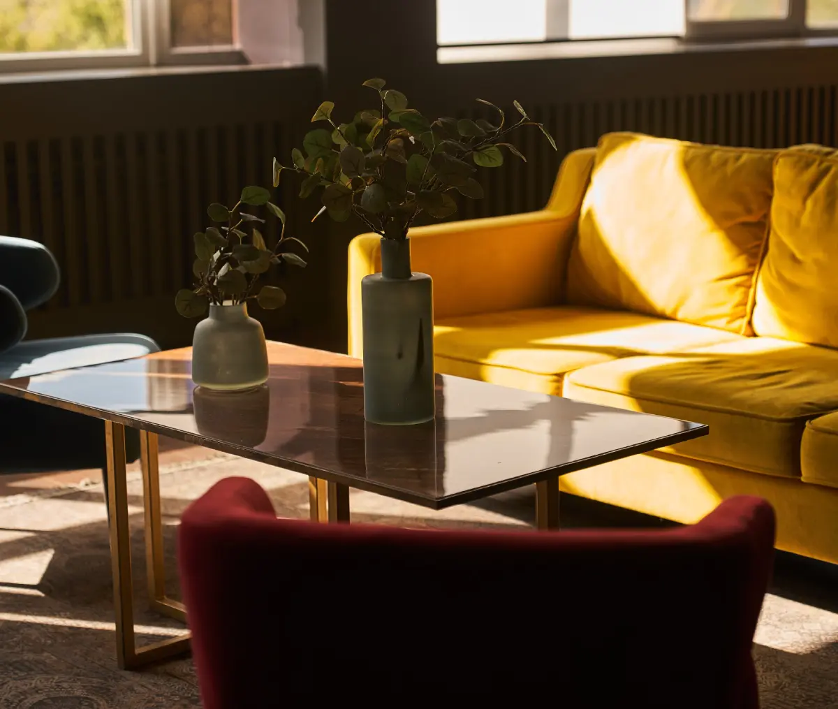 Sunlit room with a yellow sofa, red chair, and marble table. Two vases with leafy plants are on the table. Large windows in the background.