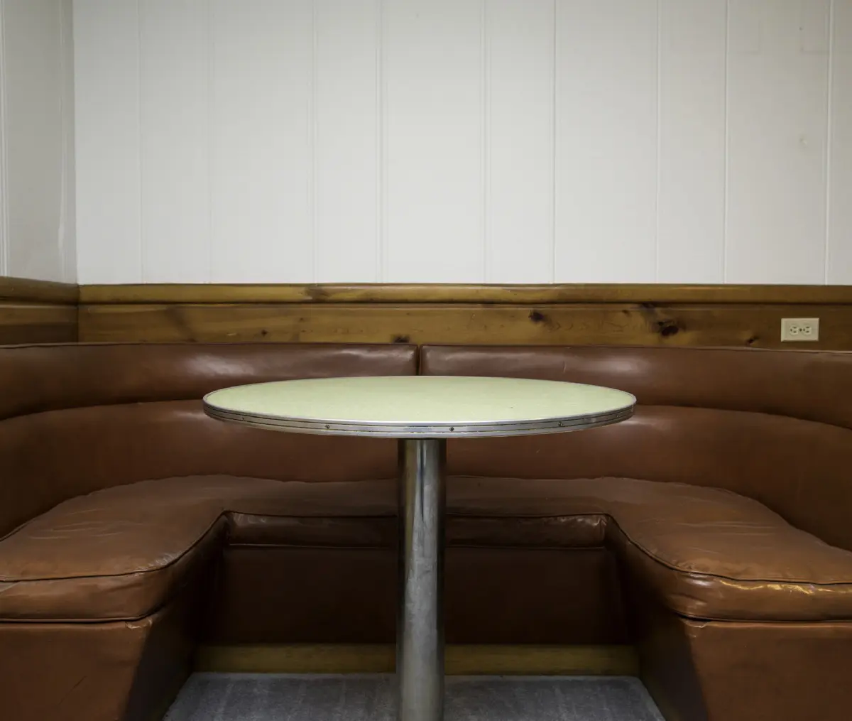 Empty round table surrounded by a curved brown leather booth in a room with wood paneling and white walls.